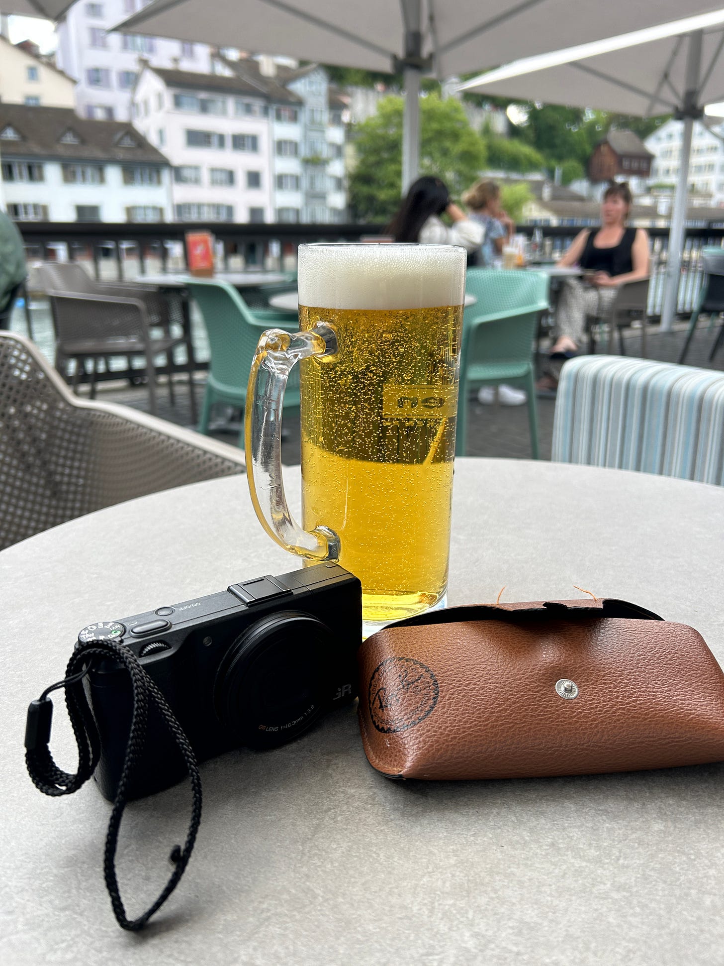 A photograph of a pint of beer on a table next to a camera and sunglasses